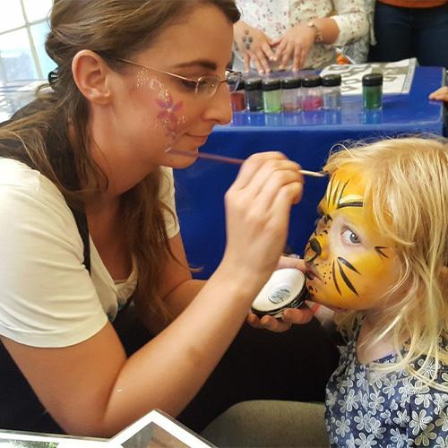 Kinderschminken Fiffikus Eine Frau mit gemalten Blumen im Gesicht malt einem Kind ein Tigergesicht