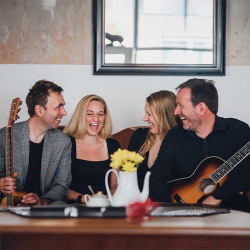 Ohrensofa Band Die Band Ohrensofa sitzt vor einem Kaffeetisch. Links sitzt ein Mann im Sacko mit einer Gitarre, daneben zwei lachende Frauen, ganz rechts noch ein lachender Mann mit Gitarre