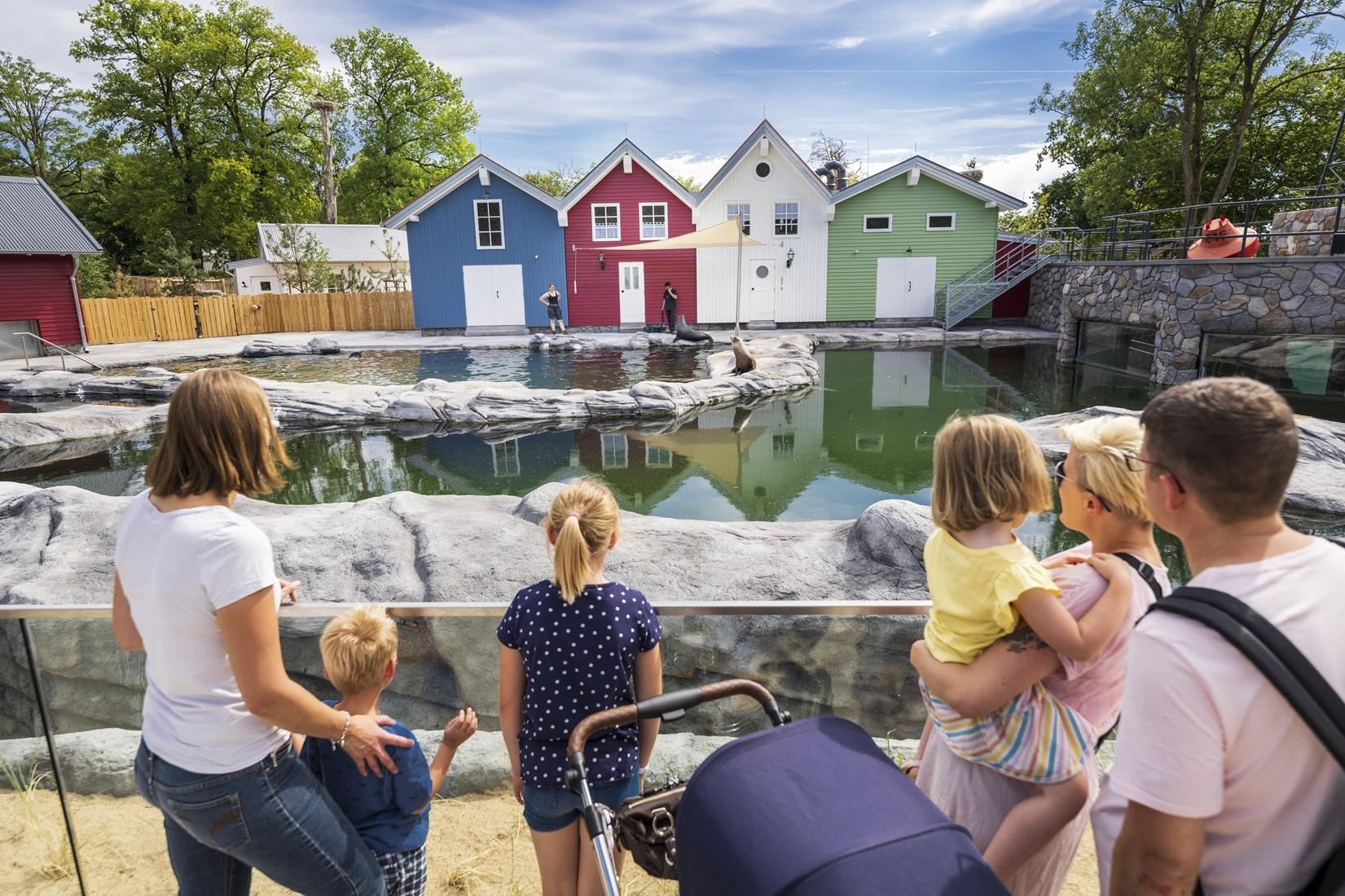 Eine Familie steht vor einem gro&szlig;en Wasserbecken. Im Hintergrund stehen bunte H&auml;user.