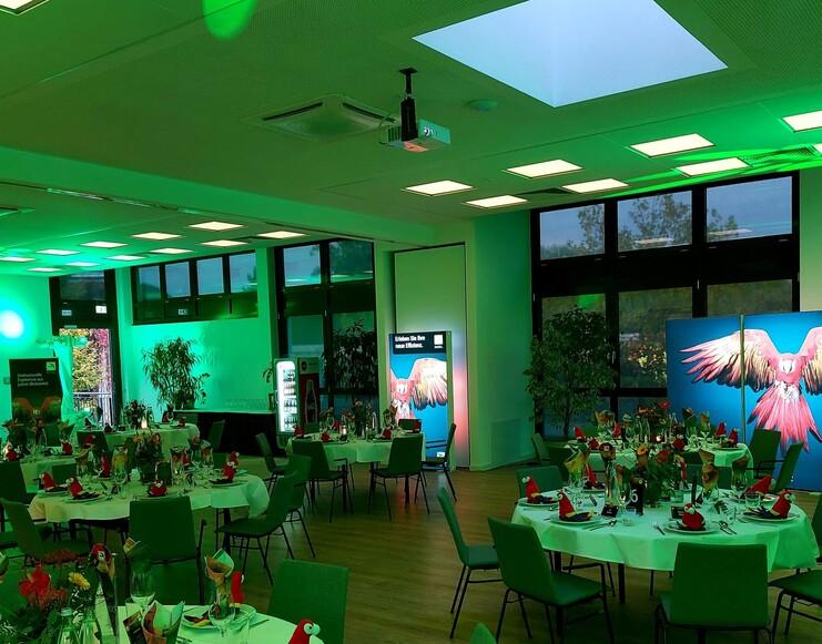 Grünes Licht Meranti-Events Ein Raum, der in grünes Licht getaucht ist. Darin stehen festlich eingedeckte, runde Tische. An den Wänden sind Bilder von roten Aras.