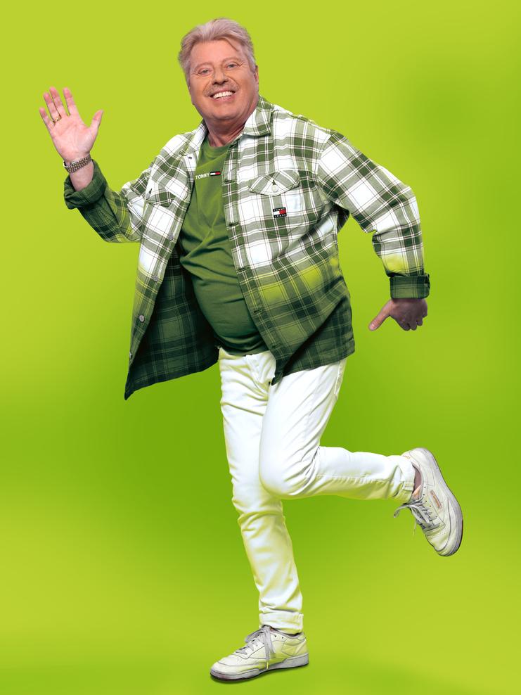 Volker Rosin Ein Mann mit weißer Hose, weißen Schuhen, grünem T-Shirt und einem weiß-grün karierten Hemd steht vor einem hellgrünen, einfarbigen Hintergrund und winkt in die Kamera. Dabei ist ein Beil angewinkelt nach hinten. Er grinst